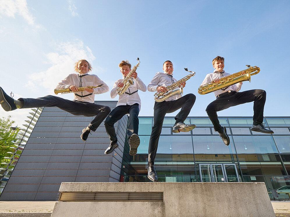 Vier Jugendliche mit je einem Saxophon in der Hand springen in die Luft.
